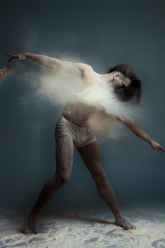 Dancing in flour concept. Long hair muscle fitness guy man male dancer in dust / fog. Guy wearing white shorts making dance element in flour cloud on isolated grey / black background © Monstar Studio
