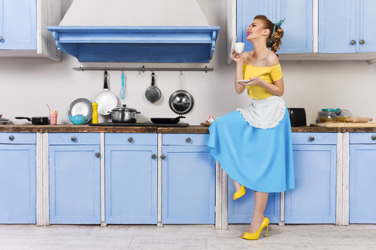 Retro pin up girl woman female housewife wearing colorful top, skirt and white apron holding and drinking cup of coffee sitting in the kitchen with utensils and tray with cupcakes and milkshake.