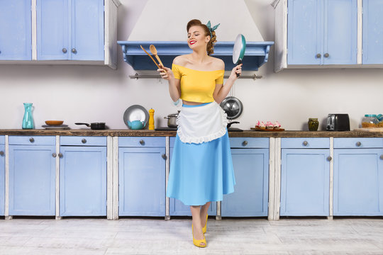 Retro pin up girl woman female housewife wearing colorful top, skirt and white apron and yellow high heels holdingwooden spoons and pan standing in the blue kitchen with blue cabinets and utensils.