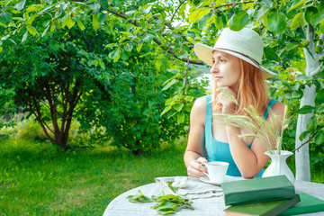 girl in a dress and a straw hat with a cup of tea, books on a table in a garden in a rural area on a summer morning, a place to work, a concept of freelancing