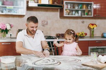 Little kid girl plays with man draws heart on scattered flour in kitchen at table. Happy family dad, child daughter cooking food cookies in weekend morning. Father's day holiday. Parenthood childhood.