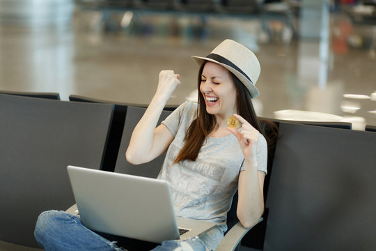 Young laughing traveler tourist woman in hat sitting working on laptop holding bitcoin doing winner gesture waiting in lobby hall at airport. Passenger traveling abroad on weekend. Air flight concept. - Powered by Adobe