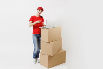 Full length portrait of delivery young woman in red cap, t-shirt isolated on white background. Female courier standing near empty cardboard boxes with laptop pc computer. Receiving package. Copy space