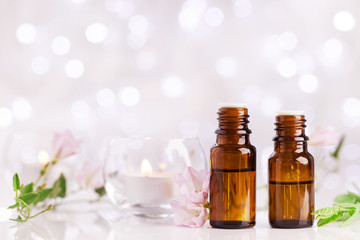 Two bottles with essential oil, flowers and candles on white table with bokeh effect. Spa, aromatherapy, wellness, beauty background.