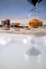 Reflection of gold sand running through the shape of modern hourglass on glowing table with currency. Time investment and passing time. Urgency countdown timer for business deadline concept