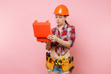 Strong young handyman woman in orange helmet, plaid shirt, denim shorts, kit tools belt full of instruments, toolbox isolated on pink background. Female in male work. Renovation and occupation concept