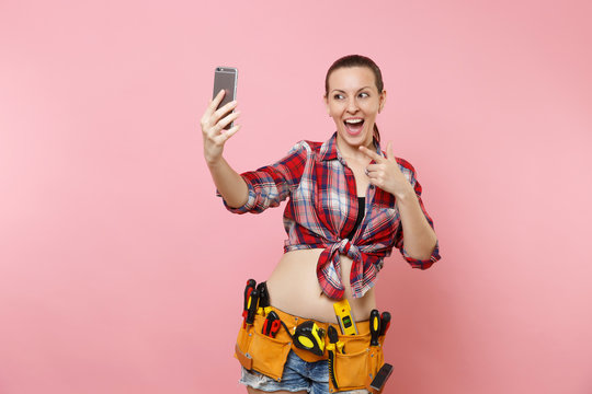 Strong excited young handyman woman in plaid shirt, denim shorts, kit tools belt full of instruments doing selfie on mobile phone isolated on pink background. Female in male work. Renovation concept.