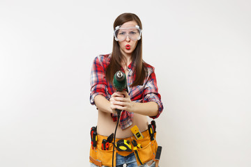 Strong young energy handyman woman in plaid top shirt denim shorts kit tools belt full of instruments in protective goggles hold power electric drill isolated on white background. Female in male work.