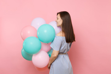 Back rear view of beautiful tender young happy woman in blue dress hold colorful air balloons looking aside isolated on bright pink background. Birthday holiday party, people sincere emotions concept.