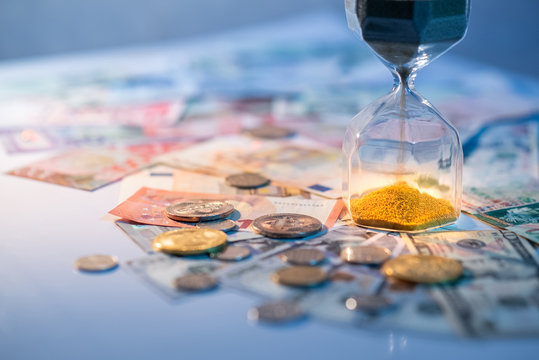 Sand running through the shape of hourglass on table with banknotes and coins of international currency. Time investment and retirement saving. Urgency countdown timer for business deadline concept
