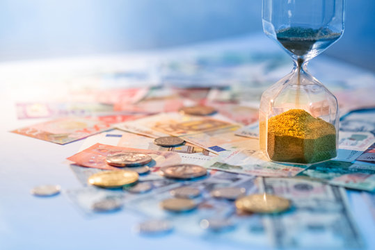 Sand running through the shape of hourglass on table with banknotes and coins of international currency. Time investment and retirement saving. Urgency countdown timer for business deadline concept