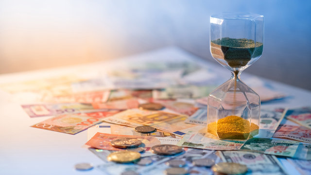 Sand running through the shape of hourglass on table with banknotes and coins of international currency. Time investment and retirement saving. Urgency countdown timer for business deadline concept