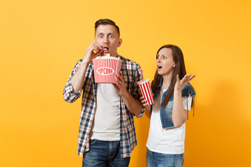 Young shocked scared couple woman man watching movie film on date eat popcorn from bucket holding plastic cup of soda or cola spreading hands isolated on yellow background. Emotions in cinema concept.