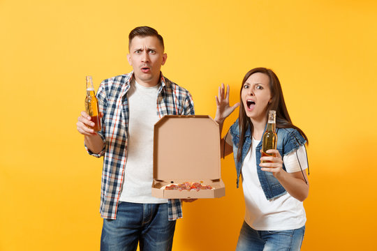 Young irritated couple woman man sport fans cheer up support team holding beer bottle italian pizza in cardboard flatbox spreading hands isolated on yellow background. Sport family leisure lifestyle.