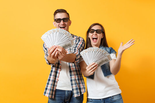 Young overjoyed couple woman and man in 3d glasses watching movie film on date holding bundle lots of dollars cash money spreading hands isolated on yellow background. Emotions in cinema concept.