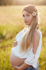 Fototapeta na wymiar Vertical shot of pregnant female keeps hand on tummy, anticipates baby, has long hair, walks across green field, looks directly at camera. Future mother with appealing look. Maternity concept