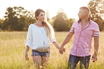 Happy young family hold hands and walk across summer field, look at each other with love. Pregnant female spends free time with husband, breath fresh air. Romance, love, relationship concept