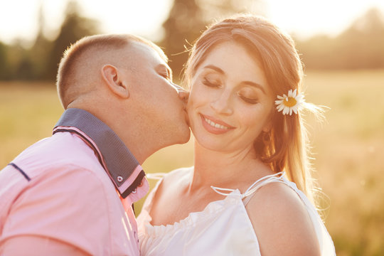 Happy young female recieves kiss from boyfriend, have outdoor walk across field, show love to each other. Lovely couple pose outside together. People, romantic relationship, togetherness concept.