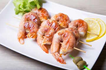 Shrimps grill in prosciutto with lemon and olives on a white plate. Close-up, selective focus.