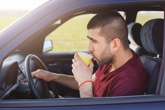 Sideways shot of attractive male driver stops to have break after driving, drinks hot coffee, looks thoughtfully on road while sits at driver`s seat. People, transportation, lifestyle, travelling