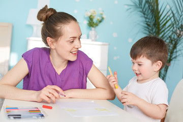 Cheerful mum and son paint together, sit at table, like creativity, spend free time at home, use...