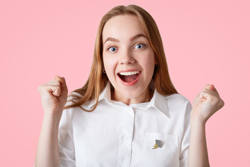 Happy young female student enjoys success, opens mouth widely and clenches fists, makes triumph gesture, stands alone against pink background, wears elegant white shirt. Peope and happiness.