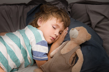 Little preschool kid boy sleeping in bed with a toy a bear.