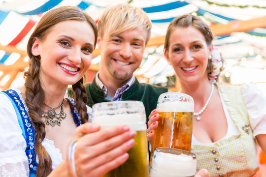Three friends at Bavarian folk fair clinking beer glasses in tent