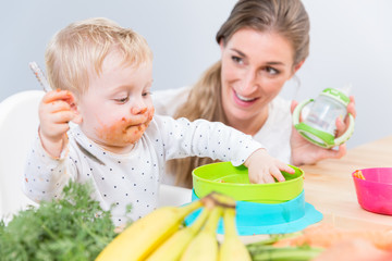 Portrait of a cute and funny baby girl sitting on high chair, while eating with her hands solid food from a plastic bowl next to her dedicated mother at home