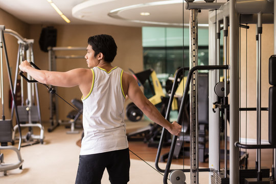 Rear view of a strong young man lifting cable with single arm, while exercising lateral or side raise for shoulder muscles in a modern fitness club