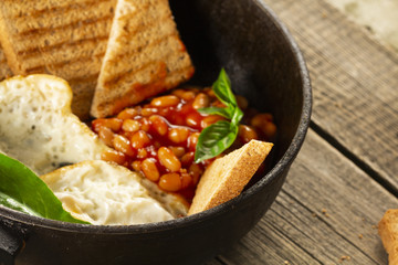 English Breakfast in cooking pan with fried eggs, sausages, bacon, beans, toasts and coffee on dark stone background