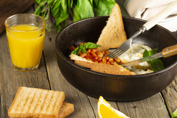 English Breakfast in cooking pan with fried eggs, sausages, bacon, beans, toasts and coffee on dark stone background