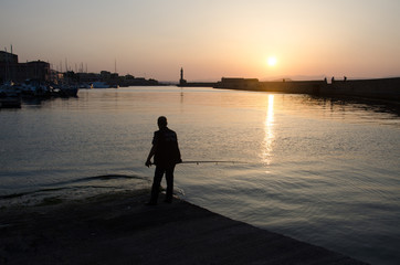 Fototapeta premium Silhouette of fisherman with lighthouse and sunset on background. Sea sunset, sunset on the sea in Greece, Chania. Golden orange sunset