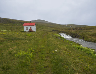 Fototapeta na wymiar Footpath in abandoned village Heysteri in Iceland West fjords in remote nature reserve Hornstrandir, small white houses with red roof, lush green grass river with water cascade and moody sky