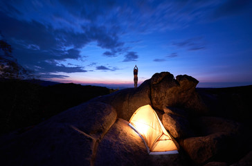 Camping on rock formation. Brightly lit tourist tent and silhouette of slim tourist girl standing with raised arms on mountain top against dark blue sky at sunset. Sport, tourism, traveling concept.