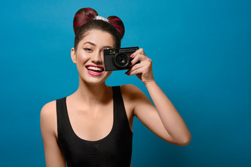 Smiling, happy girl taking photo with old camera. Laughing, keeping camera, having fancy hairstyle with a bow, colorful day make up with pink lipstick. Posing on saturated deep blue background.