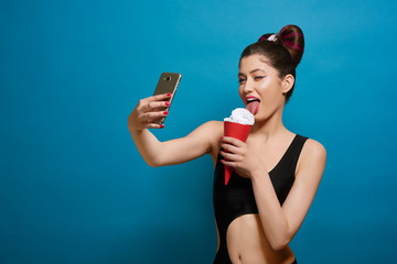 Pretty girl eating whipped cream from red paper can, taking selfie. Wearing stylish, fashionable hairstyle, colorful make up, black swimwear. Posing on saturated blue background. Contrast photo.
