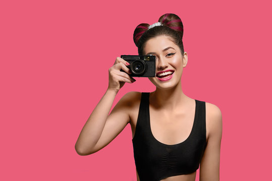 Front view of smiling, happy girl taking photo with old camera. Laughing, keeping camera, having fancy hairstyle with a bow, colorful day make up with pink lipstick. Posing on bright pink background.