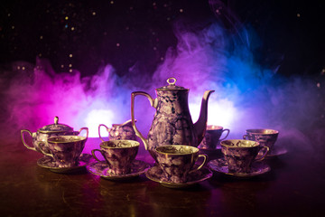 Food background tea and coffee theme. Old vintage ceramic tea or coffee pot with cups jug and sugar cup on dark background with lights and smoke. Silhouettes of ceramic tea pot and cup.