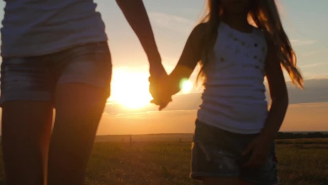 Mother and daughter walking and holding hands in the meadow at sunset. Close up shot of mother hands taking the hands of daughter at golden field on the sunset background.