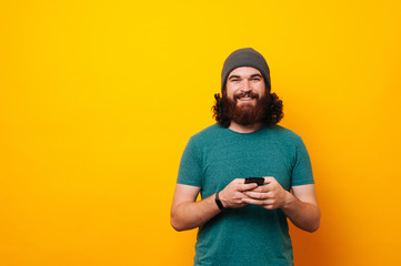 Fototapeta premium Cheerful bearded hipster man in t-shirt holding phone and looking at the camera over yellow background with copyspace
