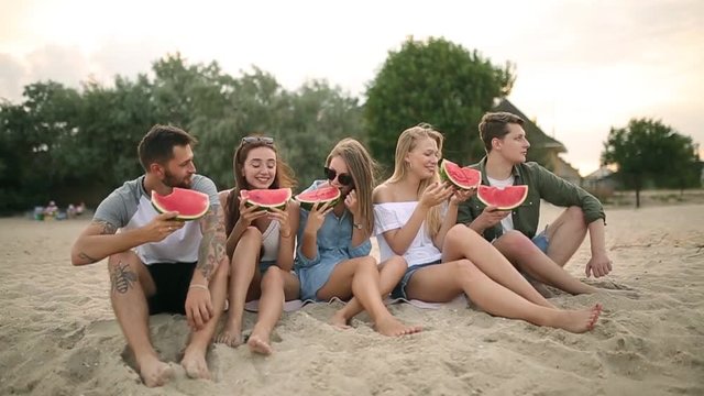 Happy Friends Eating Watermelon Sitting on Sandy Beach on Vacation. Young Men and Women Wearing Blue Jeans Shorts. Friendship and Summer Concept