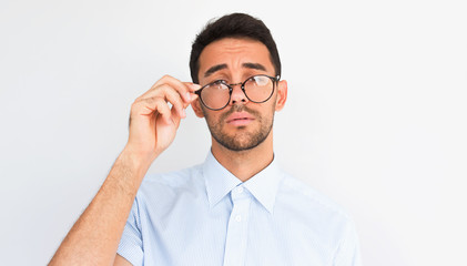 Closeup image of handsome bearded young male looking to the camera, touches rim of round spectacles, wears blue shirt, stands against studio background. People, business, health, emotions concept
