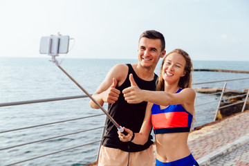 Joyful sportswoman and sportsman in sportswear showing a thumb up while taking a selfie on smartphone, after workout outdoors, near the sea.