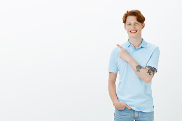 Time to share positive emotions. Positive handsome redhead male in trendy clothes with tattoed arm, pointing at upper left corner and smiling broadly, suggesting great place to watch film together