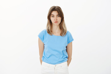 Fototapeta premium Portrait of unhappy cute female coworker in blue t-shirt, pouting and sulking, gazing at camera with lack of interest, being indifferent and unimpressed, standing upset over gray background
