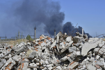 A pile of concrete rubble with protruding rebar on the background of thick black smoke in the blue sky.