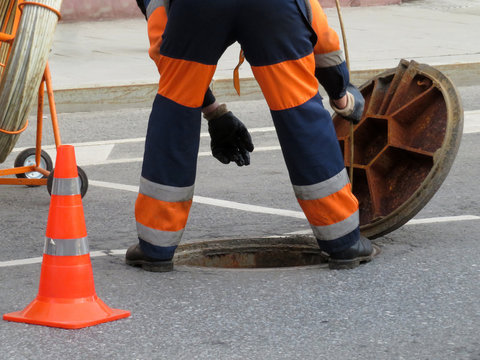 Worker In Uniform Stand Over The Open Sewer Hatch. Repair Of Sewage Or Underground Utilities, Nightman Cleans Drains, Cable Laying