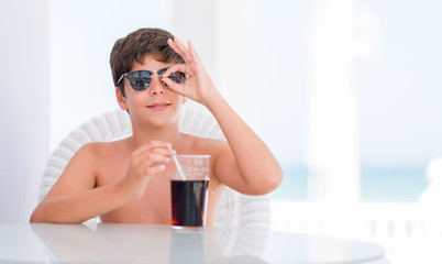 Young child on holidays drinking soda with happy face smiling doing ok sign with hand on eye looking through fingers