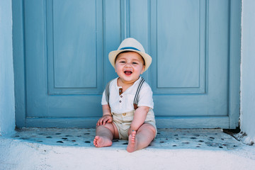 Beautiful baby sitting in front of a gray door in Santorini, Greece, wearing a hat